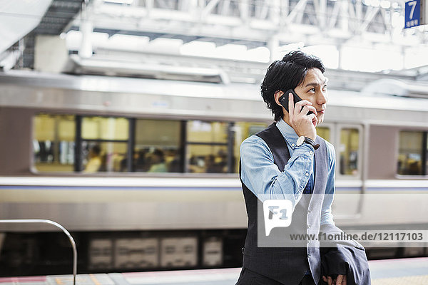 Businessman wearing blue shirt and vest standing on train station platform  talking on mobile phone.