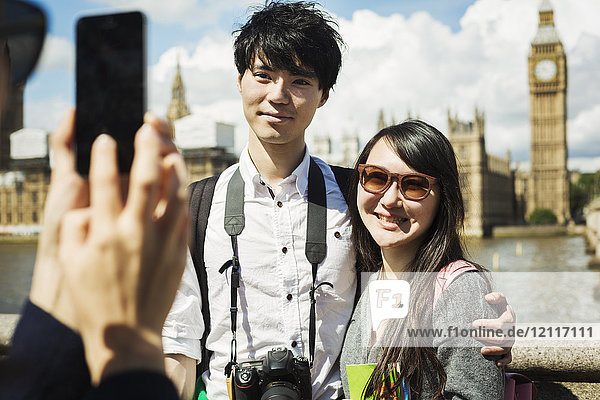 Lächelnde Frau mit schwarzen Haaren  die ein Paar mit einem Smartphone fotografiert  das auf der Westminster Bridge über die Themse in London steht  mit dem Houses of Parliament und Big Ben im Hintergrund.