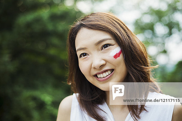 Portrait of young woman with brown hair  Japanese flag painted on her cheek  smiling at camera.