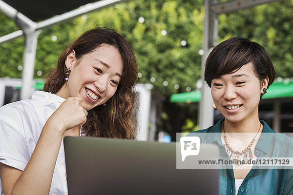 Zwei Frauen mit schwarzen Haaren in grün-weißem Hemd sitzen lachend vor einem Laptop am Tisch in einem Straßencafé.