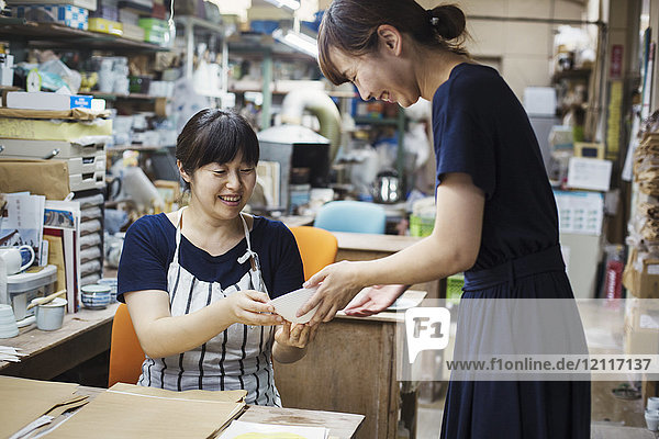 Zwei lächelnde Frauen sitzen und stehen in einer Werkstatt und schauen auf eine japanische Porzellanschüssel.