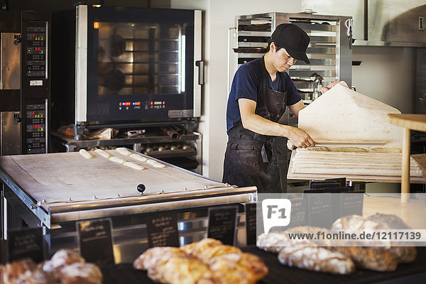 Mann  der in einer Bäckerei arbeitet  bereitet ein großes Blech mit Teig für Brötchen vor  im Hintergrund der Ofen.