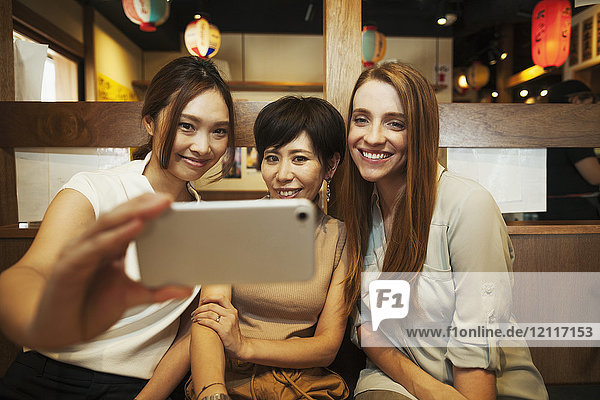 Drei Frauen sitzen nebeneinander in einem Restaurant und nehmen Selfie mit Smartphone.