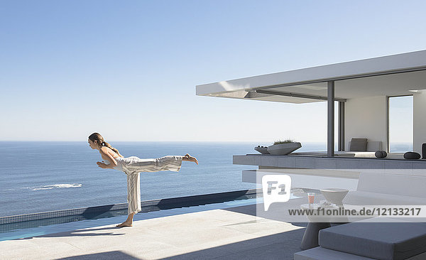 Frau übt Yoga-Krieger 3 Pose auf sonnigen modernen  luxuriösen Haus Schaufenster Außenterrasse mit Meerblick