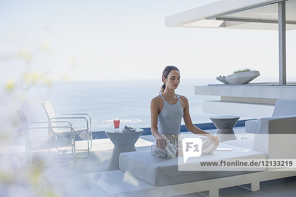 Gelassene Frau meditiert im Lotussitz auf modernem  luxuriösem Vorzeigesofa auf der Terrasse mit Meerblick