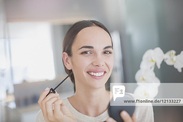 Porträt einer lächelnden  selbstbewussten Frau  die Wimperntusche mit einem Wimperntuschestift und einem Kompaktspiegel aufträgt