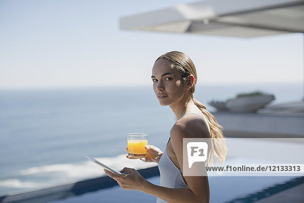 Porträt einer Frau  die ein digitales Tablet benutzt und Orangensaft auf einer sonnigen  luxuriösen Terrasse mit Meerblick trinkt