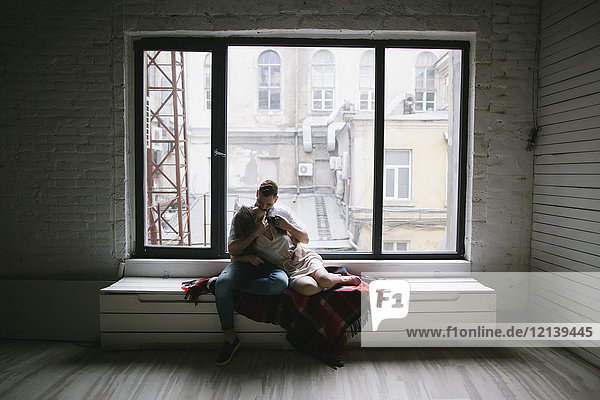 Kaukasisches Paar küsst sich auf einer Bank in der Nähe des Fensters