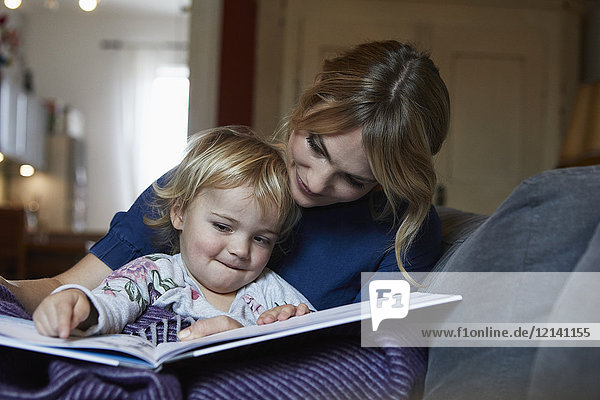 Mutter und kleine Tochter sitzen zu Hause auf der Couch und schauen sich das Buch an.