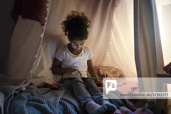 Kleines Mädchen sitzt im dunklen Kinderzimmer und schaut auf ein digitales Tablett.