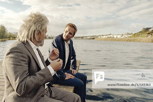 Two businessmen sitting on jetty at a lake talking