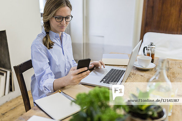 Woman using cell phone and laptop on wooden desk at home