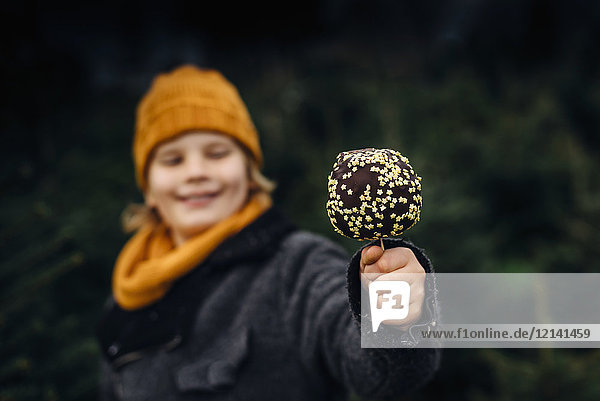 Kleiner Junge steht vor Tannenbäumen und hält einen schokoladengetauchten Apfel.