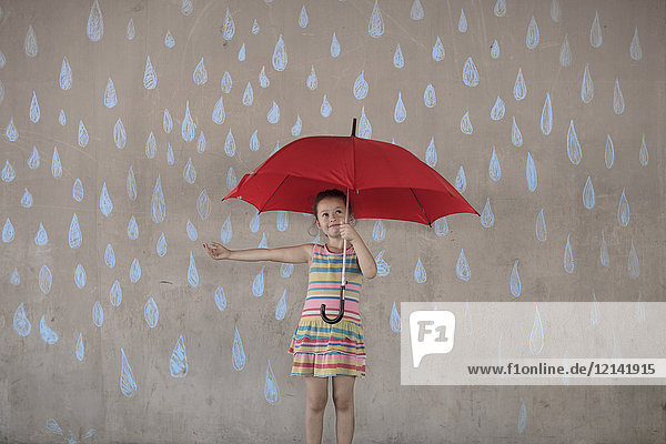 Mädchen mit einem roten Regenschirm neben einer Betonmauer mit Zeichnungen von Regentropfen.