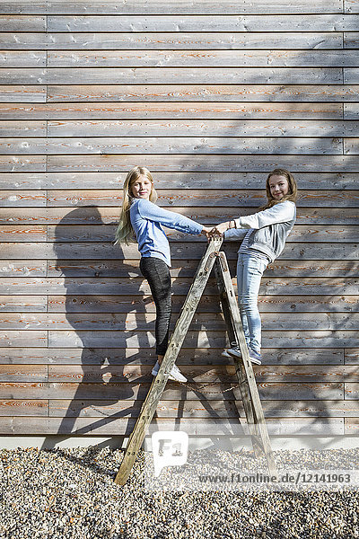 Zwei Mädchen  die auf einer Leiter vor einer Holzfassade stehen.