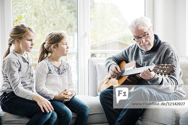 Two girls sitting on sofa listening to grandfather playing guitar