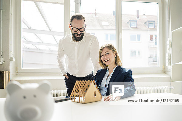Laughing woman and man with house model and piggy bank in office