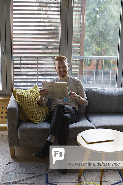 Lächelnder Mann sitzt zu Hause auf der Couch und liest Zeitung.