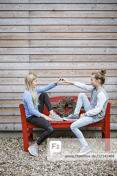 Two best friends sitting on a red bench in front of a wooden facade forming heart with their hands