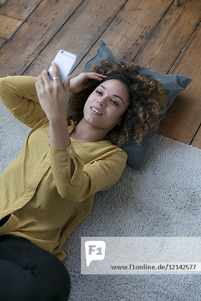Smiling young woman lying on the floor at home with cell phone