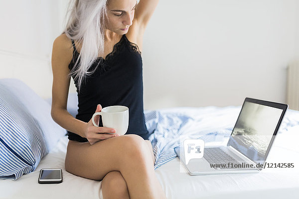 Young woman in bed with cup of coffee and laptop