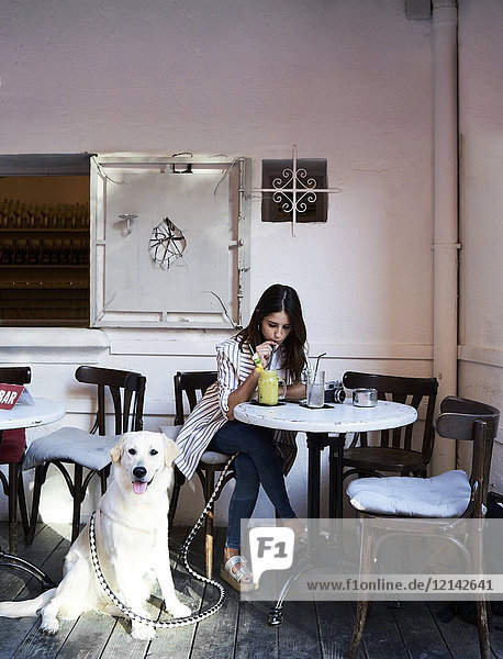 Junge Frau sitzt mit ihrem Hund im Café und trinkt einen Smoothie.