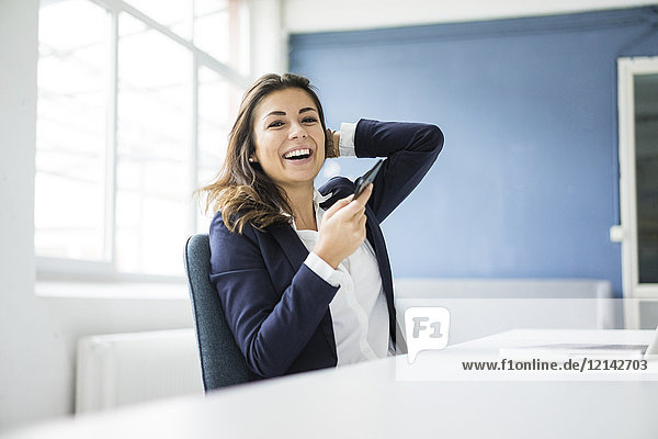 Porträt einer lachenden Geschäftsfrau mit Handy am Schreibtisch im Büro