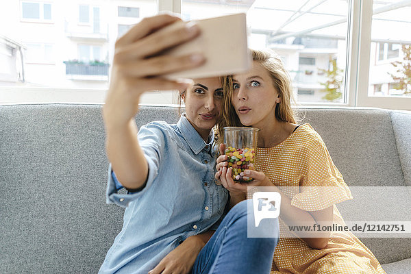 Zwei junge Frauen sitzen auf der Couch und nehmen einen Selfie mit Süßigkeiten.