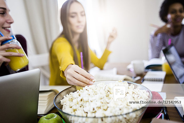 Female student with friends taking popcorn from bowl at table at home