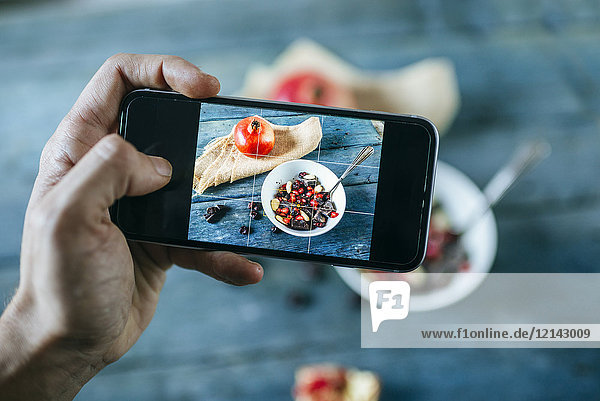 Männerhand beim Fotografieren mit Smartphone von Schale mit Dessert aus Mandeln  Granatapfel und Schokolade