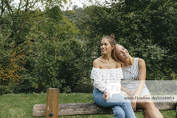 Zwei glückliche junge Frauen sitzen auf einem Zaun in einem Park.