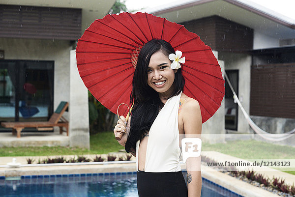 Portrait of smiling woman with flower in her hair holding a red tradtional umbrella at a swimming pool