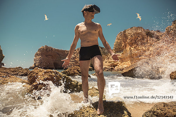 Erwachsener Mann mit VR-Brille zwischen Felsen am Strand