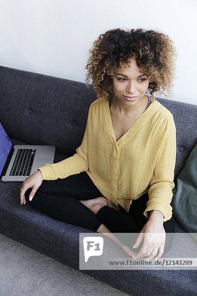 Smiling young woman sitting on couch at home next to laptop