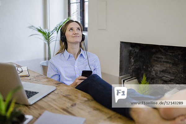 Smiling woman sitting at desk at home with feet up listening to music