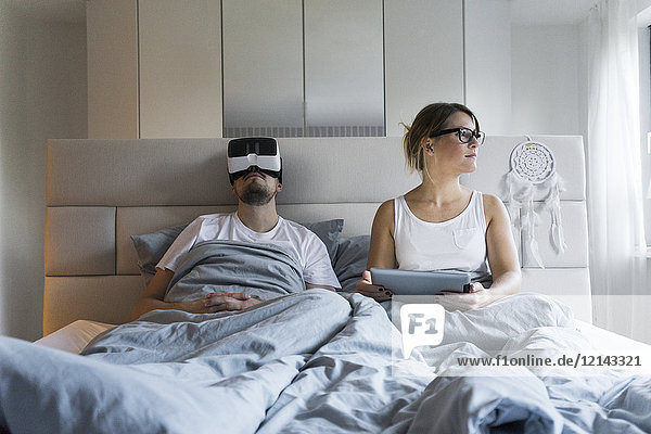 Couple in bed at home with tablet and VR glasses