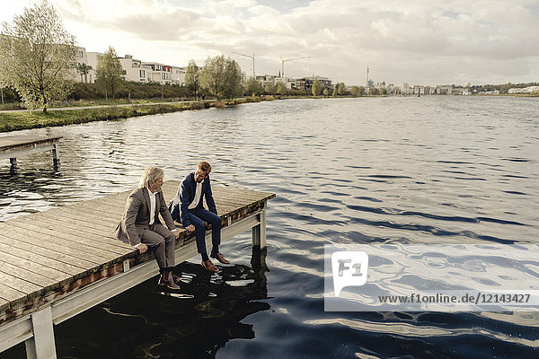 Zwei Geschäftsleute  die auf einem Steg am See sitzen und sich unterhalten.
