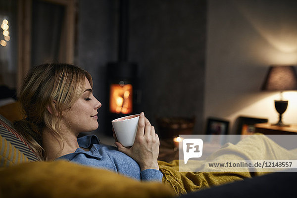 Lächelnde Frau mit einer Tasse Kaffee  die abends zu Hause auf der Couch sitzt.