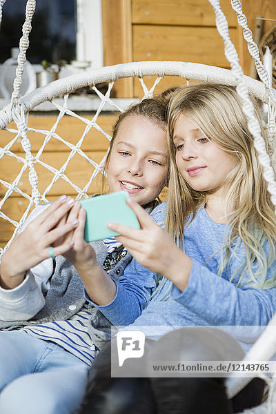 Porträt von zwei Mädchen  die sich in einem Hängestuhl entspannen und sich mit dem Smartphone selbst versorgen.