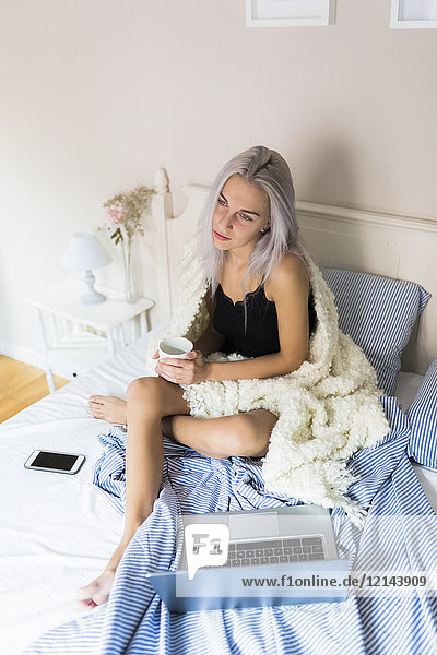 Pensive young woman in bed with cup of coffee and laptop