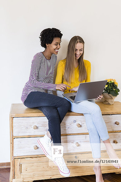 Zwei junge Frauen sitzen auf dem Schrank und schauen zusammen auf den Laptop.