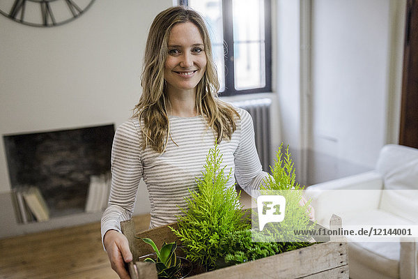 Porträt einer lächelnden Frau  die eine Kiste mit Pflanzen zu Hause trägt.