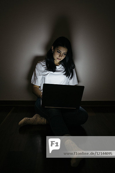 Junge Frau zu Hause auf dem Boden sitzend mit Laptop im Dunkeln