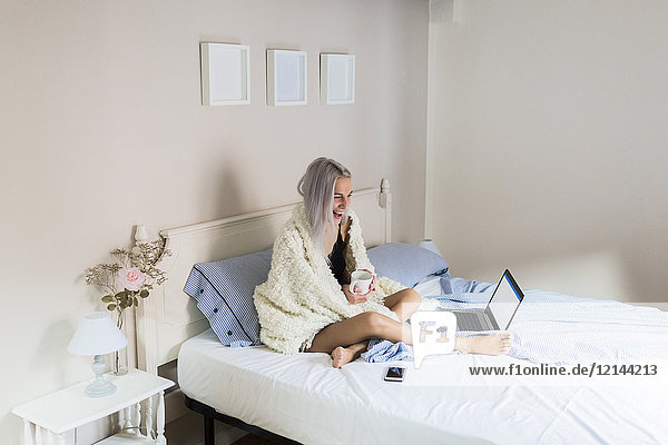 Glückliche junge Frau im Bett mit Tasse Kaffee und Laptop