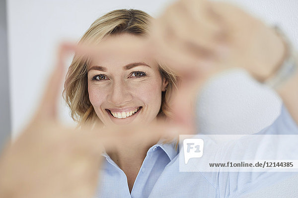 Porträt einer lächelnden  blonden Frau  die mit den Fingern den Rahmen baut  während sie den Betrachter ansieht.