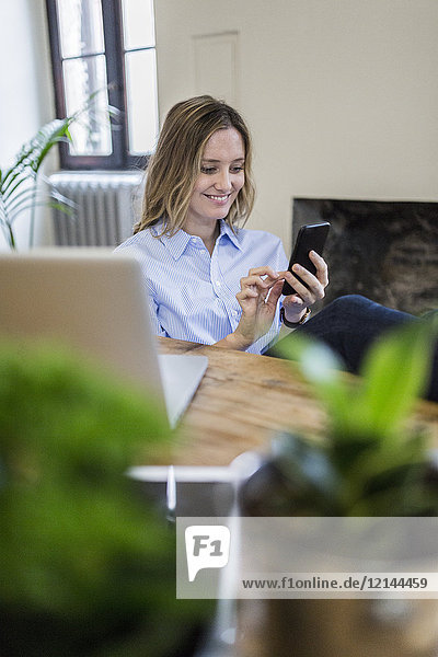 Lächelnde Frau sitzt zu Hause am Schreibtisch und überprüft das Handy
