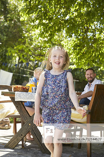 Porträt eines lachenden Mädchens mit Eltern im Hintergrund am Gartentisch
