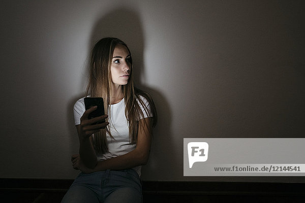 Junge Frau zu Hause auf dem Boden sitzend mit dem Handy im Dunkeln