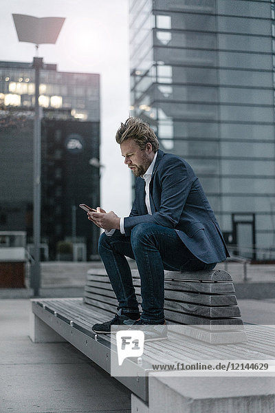 Businessman sitting on bench in the city using cell phone