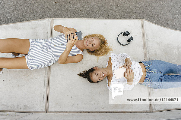 Zwei glückliche junge Frauen mit Handys auf einer Rampe in einem Skatepark.